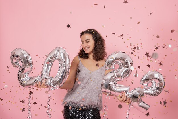 Linda menina morena com cabelos cacheados e roupas festivas, posando em um fundo rosa com confete e segurando balões de prata dos números 2022.