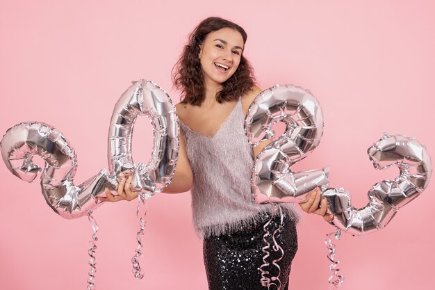 Linda menina morena com cabelos cacheados e roupas festivas, posando em um fundo rosa com confete e segurando balões de prata dos números 2023.