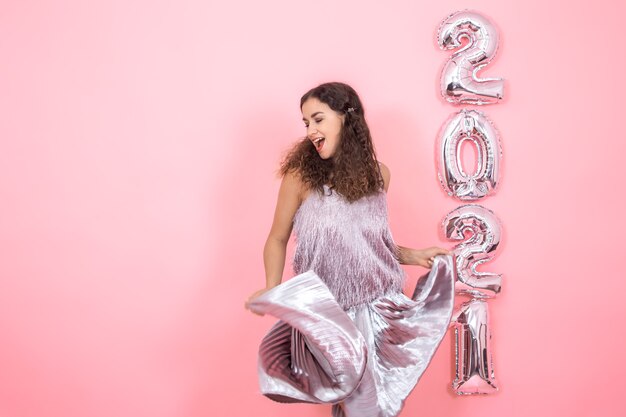 Linda menina morena com cabelos cacheados em roupas festivas em movimento em uma parede rosa com balões de prata para o conceito de ano novo