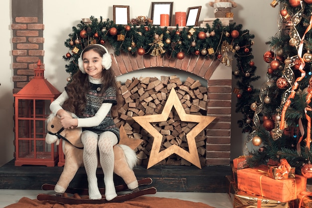 Linda menina morena com cabelos longos, sentado em um cavalo de brinquedo na sala de Natal decorada.