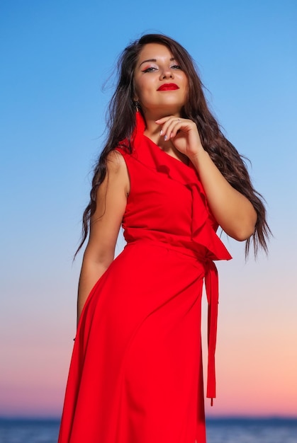 Linda menina morena com um vestido vermelho, posando à beira-mar durante o nascer ou o pôr do sol