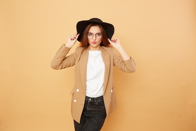 Linda menina morena elegante com cabelo comprido em óculos e um chapéu preto na cabeça, vestida de camiseta branca, jeans e jaqueta bege está posando sobre o fundo bege no estúdio.