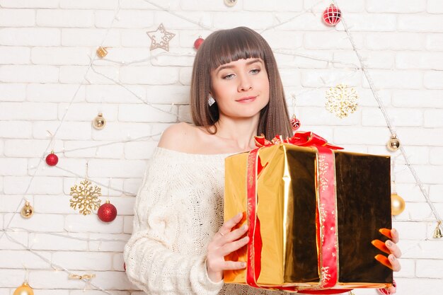 Linda menina morena encaracolada para a árvore de Natal com brinquedos, decorações, luzes. Mulher jovem com um suéter de malha quente, legging longa de golfe segurando uma caixa de presente nas mãos, sorrindo junto à lareira