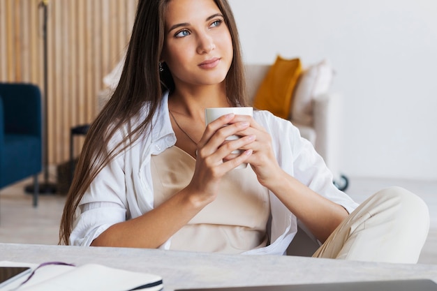 Foto linda menina morena está sentada à mesa em frente ao laptop, segurando uma xícara de café branca com as mãos.