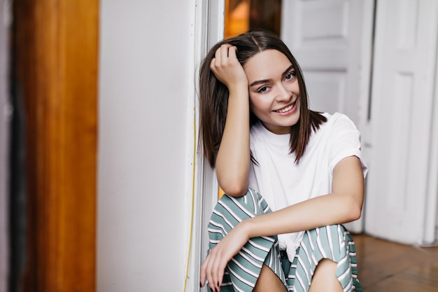 Linda menina morena posando no chão Retrato interior de uma agradável modelo feminina tocando seu cabelo