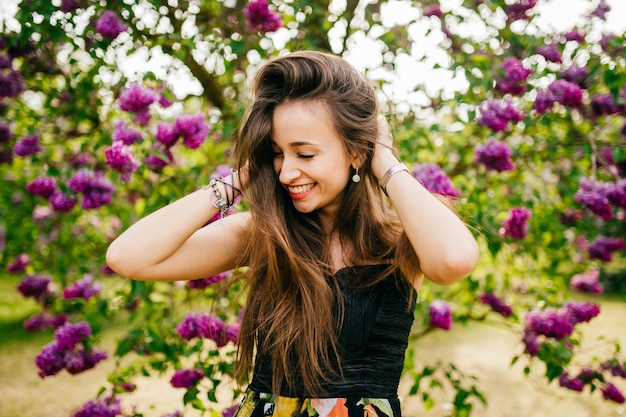 Linda menina morena segurando seus longos cabelos e sorrindo sobre a árvore de florescência lilás