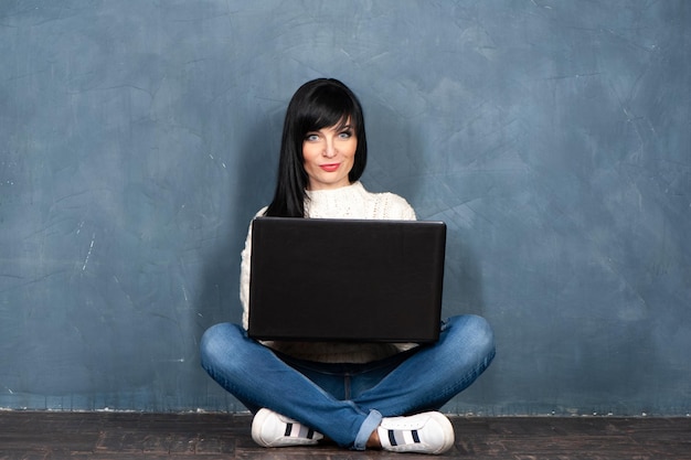 Linda menina morena senta-se no chão com seu laptop olha para a câmera e sorri Retrato de estúdio em um fundo gasto na moda azul monocromático