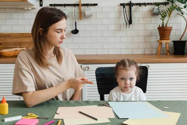 Linda menina pré-escolar com raiva fazendo lição de casa criativa com sua mãe enquanto está sentado à mesa