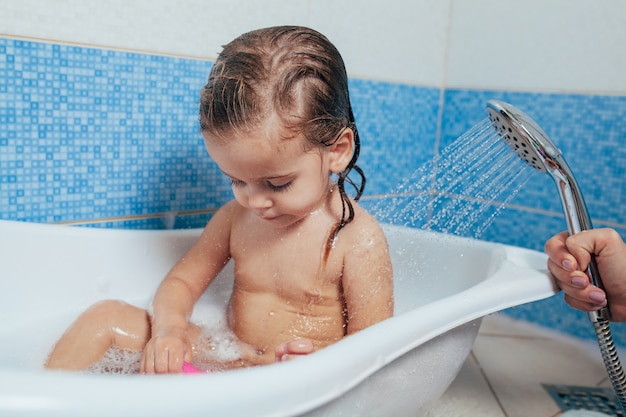 Linda menina tomando banho em casa. Um bebê fofo está sentado no banheiro e brincando com brinquedos e água.