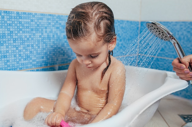 Linda menina tomando banho em casa. um bebê fofo está sentado no banheiro e brincando com brinquedos e água.