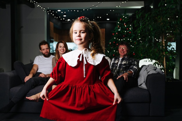 Linda menina vestida de vermelho, posando para uma foto. Sua grande família sentada em um sofá no fundo junto com a árvore de Natal.