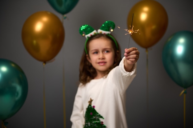 Linda menina vestindo uma argola de duende e um suéter branco aconchegante com glitter, uma árvore de Natal que fica em frente a balões de ar inflados verdes dourados e ilumina os brilhos que comemoram o ano novo de 2022