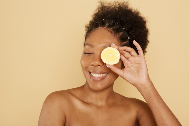 Linda modelo feminina afro-americana segurando meio limão na frente do rosto em um fundo bege o conceito de cuidar de cosméticos com vitamina c cuidados com a pele e spa