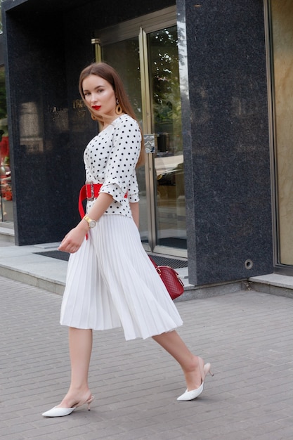 Linda modelo parece mulher morena com vestido branco com bolinhas pretas é enquanto acorda em um plano de fundo de rua da cidade com bolsas elegantes na mão.