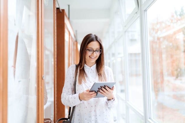 Linda morena caucasiana em pé ao lado do noticeboard e usando o tablet em pé no prédio da Universidade.