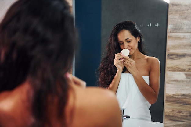 Linda morena em pé no banheiro perto do espelho e limpando o rosto.