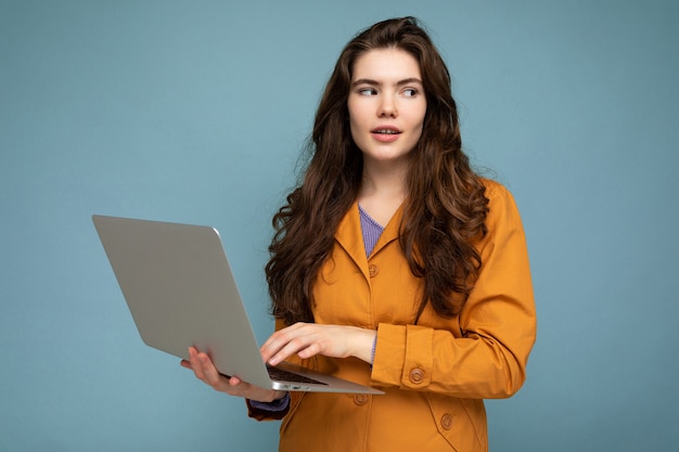 Linda morena encaracolada jovem segurando o computador netbook, olhando para o lado vestindo uma jaqueta amarela, digitando no teclado isolado sobre fundo azul.