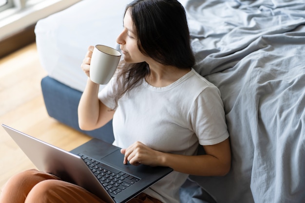 Linda morena jovem trabalhando em um laptop e bebendo café, sentada no chão perto da cama pela janela panorâmica, com uma bela vista do andar alto. Interior moderno e elegante