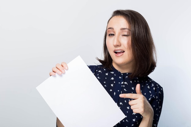 Linda morena mostra um dedo em um papel branco limpo, pisca com um sorriso, felicidade, alegria, emoções