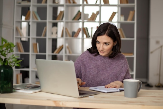 una linda morena con un suéter lila está sentada en una mesa con una laptop tomando notas