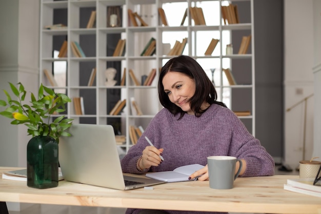 una linda morena con un suéter lila está sentada en una mesa con una laptop tomando notas