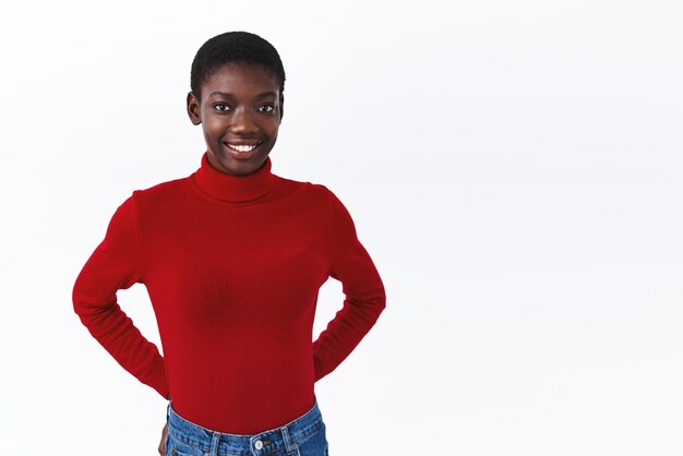 Linda mujer afroamericana tímida con corte de pelo corto en cuello alto rojo con las manos detrás