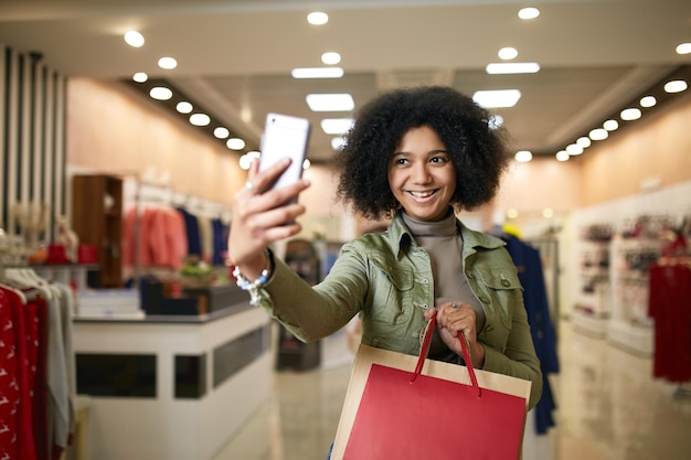 Linda mujer afroamericana tomando selfie con bolsas de compras y sonriendo cerca de la tienda de ropa negra