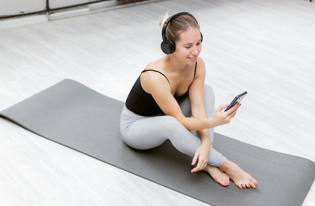 Linda mujer alegre en ropa deportiva escuchando música con auriculares y usa un teléfono inteligente mientras se acuesta en el interior de la alfombra