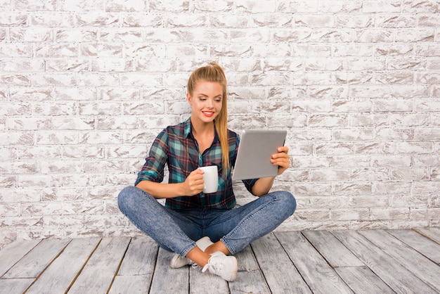 Linda mujer alegre con tableta y taza sentada en el floar