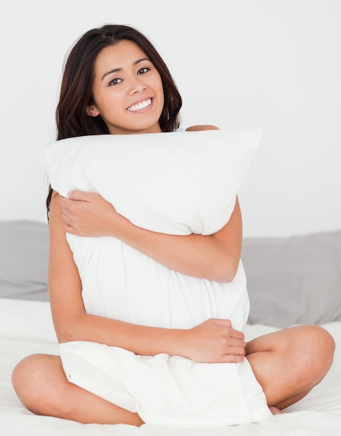 Linda mujer con almohada sentada en su cama sonriendo a la cámara