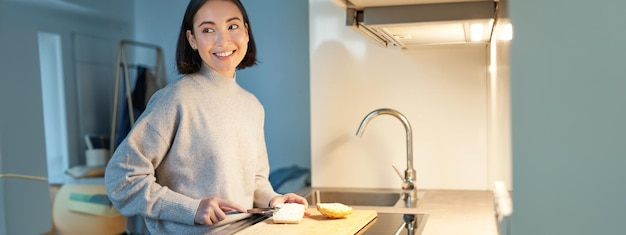 Linda mujer asiática haciéndose tostadas y cortando una barra de pan preparando sándwich en la cocina