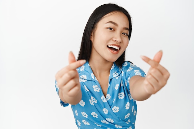 Linda mujer asiática mostrando los dedos del corazón y sonriendo a la cámara con un vestido azul de pie sobre fondo blanco.