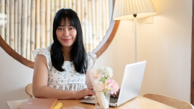Una linda mujer asiática se sienta en una mesa en una cafetería con su computadora portátil sonriendo a la cámara