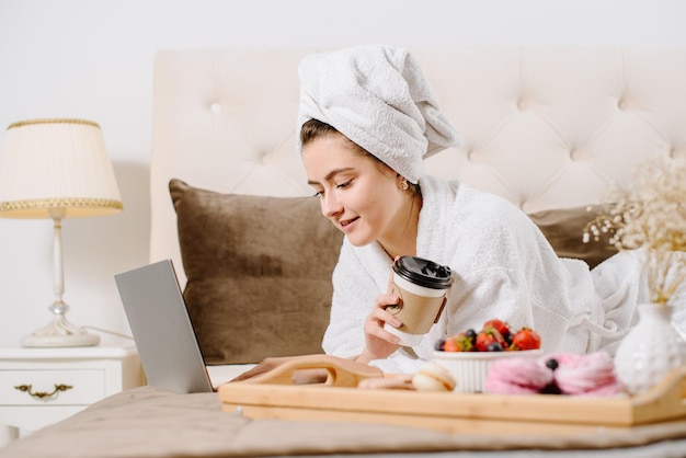 Linda mujer en bata de baño y toalla en la cabeza usando una computadora portátil en la cama en casa