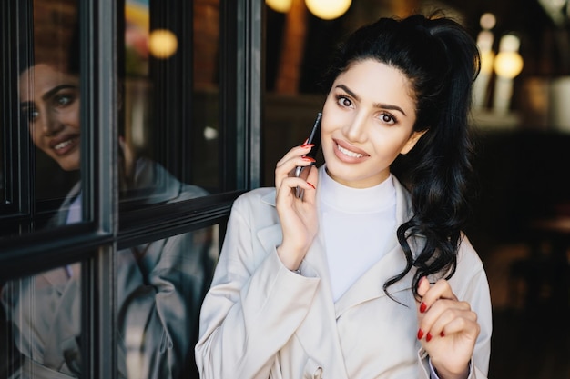 Linda mujer con cabello oscuro ondulado, ojos encantadores, labios bien formados y dientes blancos y uniformes, vestida de blanco, comunicándose por teléfono tocando suavemente su cabello