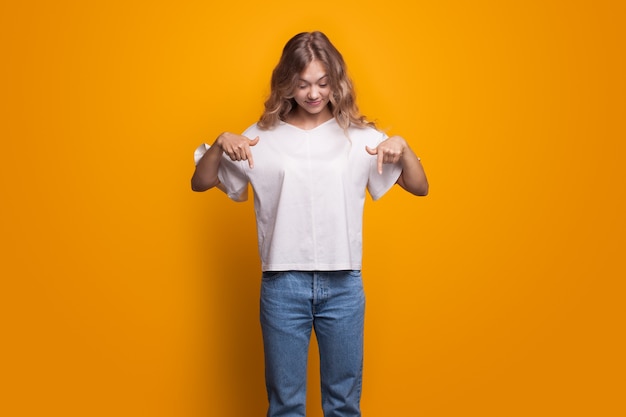 Linda mujer con cabello rizado rubio apuntando hacia abajo con una camiseta blanca y jeans posando en una pared amarilla