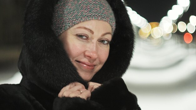 Linda mujer caminando por el abrigo de piel negro Chica al aire libre camina en una noche de invierno entre las luces de la calle de la ciudad Parque de nieve con guirnaldas Hermosa sonrisa feliz sensual