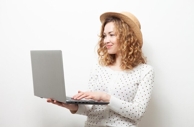 Una linda mujer caucásica de pelo rizado sonriente con sombrero de paja usa una laptop con fondo blanco