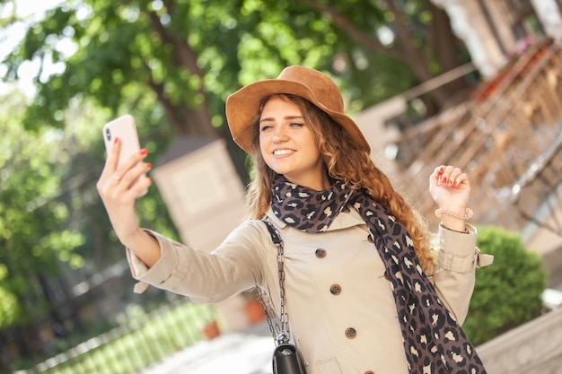 Linda mujer caucásica sonriente con sombrero y gabardina hace selfie en la ciudad