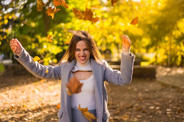 Linda mujer disfrutando del otoño en un parque al atardecer tirando hojas de los árboles