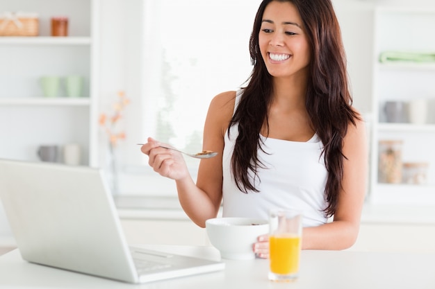 Linda mujer disfrutando de un tazón de cereales mientras se relaja con su computadora portátil