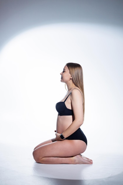 Linda mujer embarazada con cabello largo en ropa interior negra está sentada sobre un fondo blanco