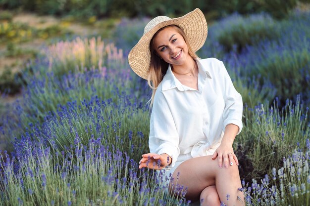 Linda mujer feliz disfrutando de la naturaleza en el prado de lavanda La mujer joven se sienta en camisa blanca y sombrero