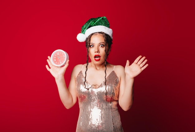 Una linda mujer feliz con un sombrero de Papá Noel sostiene un despertador sobre un fondo rojo El concepto de la temporada navideña Navidad y año nuevo