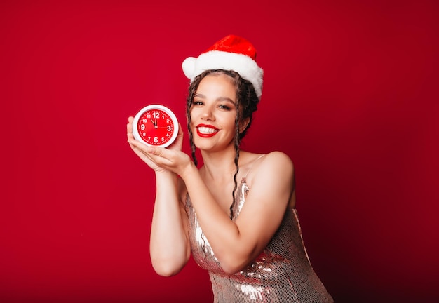 Una linda mujer feliz con un sombrero de Papá Noel sostiene un despertador sobre un fondo rojo El concepto de la temporada navideña Navidad y año nuevo