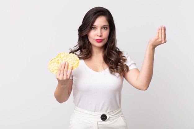 Linda mujer haciendo capice o gesto de dinero diciéndote que pagues y sosteniendo una dieta pasteles de arroz