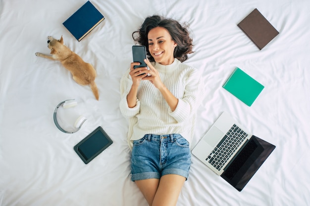 Linda mujer hermosa joven feliz está acostado en la cama en casa con su perrito y algunas cosas para estudiar. Chica con laptop y teléfono