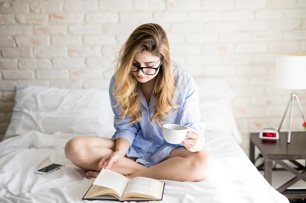 Linda mujer inteligente con gafas leyendo un libro y bebiendo café por la mañana