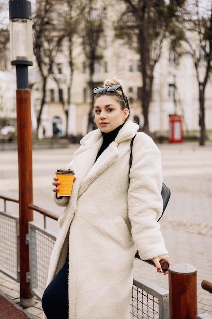 Una linda mujer joven con un abrigo de piel blanco y una taza de café camina por la ciudad Elegante joven embarazada Moda informal para mujeres