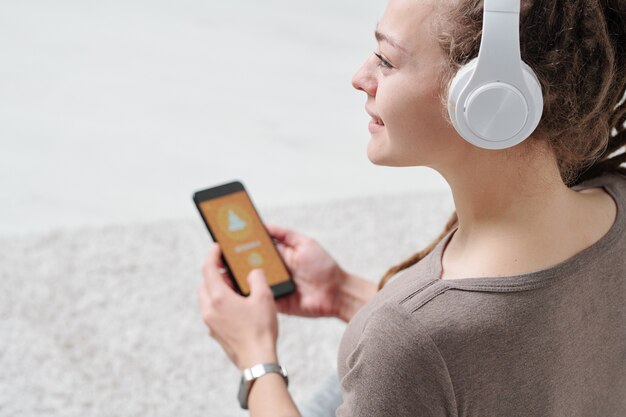 Foto linda mujer joven en auriculares y ropa deportiva sosteniendo un teléfono inteligente mientras escucha lecciones de audio de la práctica de yoga